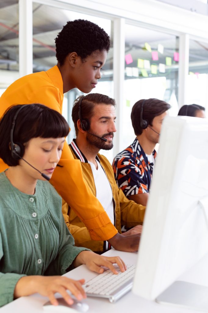 Side view of diverse customer service executive trainer assisting her team at desk in office